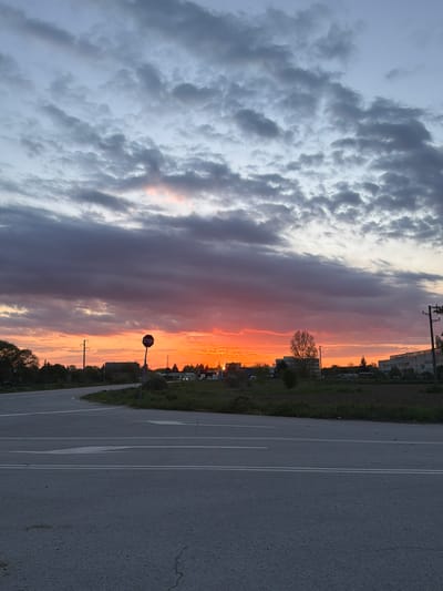 The image depicts a sunset scene in what appears to be a parking lot or paved area, possibly located in or near Dolni Chiflik, Bulgaria. The sky is divided, with the left side showing overcast, gray clouds, while the right side showcases vibrant orange and red hues of a setting sun. In the middle ground, a "STOP" sign is visible next to some greenery. Distant buildings and trees are silhouetted against the sunset colors. The foreground comprises a paved surface with white lane markings. There are no people visible in the image.