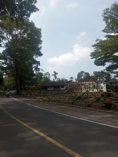 A daytime exterior scene in Niz Lakuk, India, presents an asphalt road with a solid yellow centerline and a broken white shoulder line. A person on a light-colored motorcycle is observed traveling away from the camera on the left lane. To the right of the road, a low, rustic fence made of bamboo or slender wooden poles demarcates a property. Behind this fence, dark, uneven ground leads to two structures. The first structure, positioned to the left, is a single-story building featuring a dark tiled roof and light-colored walls, displaying a partially visible "Shinfa" sign. Adjacent to it on the right, a second structure is undergoing construction or renovation, evidenced by extensive wooden scaffolding encasing a partially formed building. A solitary individual wearing an orange or red shirt is visible near the fence line on the right side of the construction zone. The environment is heavily vegetated, with numerous large trees featuring dense green foliage lining the road's left edge and forming a natural backdrop across the horizon. The sky is clear, light blue with scattered white cumulus clouds.