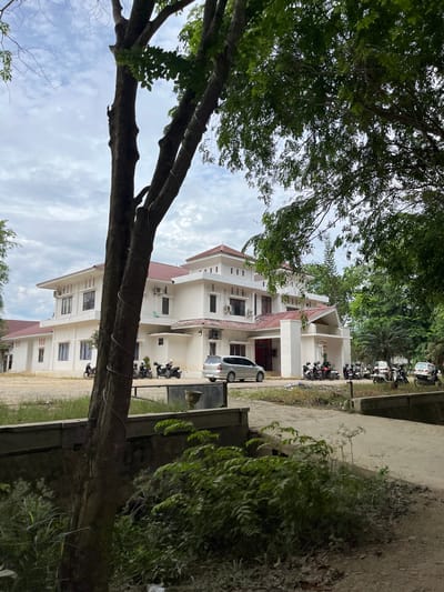 The image depicts the exterior of a white, multi-story building with a red tiled roof, located in Bundar, Indonesia. The building has multiple windows and visible air conditioning units. In front of the building, several motorcycles are parked, some with helmets on them, suggesting recent or ongoing activity. A silver car is parked on the right side of the image, and another vehicle is visible further back.
The foreground of the image is dominated by the trunks of two large trees, with one tree's branches and leaves obscuring parts of the building and the sky. The sky is overcast with clouds. The ground in front of the building appears to be a paved area, with a dirt path or road leading into the scene. A low concrete wall runs horizontally across the middle ground, separating the building's vicinity from a vegetated area. Lush green foliage is present in the lower right and center foreground. The overall environment appears to be semi-urban or a campus-like setting, with a mix of built structures and natural elements.