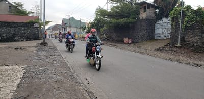 The image shows a street scene in Keshero, Democratic Republic of the Congo. There are three motorcyclists traveling on an asphalt road, which is bordered on both sides by a rock wall. The road is in poor condition, with areas of uneven pavement and loose gravel. The motorcyclists are wearing helmets and are all traveling in the same direction. There are a few small buildings and trees on the sides of the road. The motorcyclists are traveling on the right side of the road. One of the motorcyclists is wearing a green shirt with the word “STRATEGIC” on it. There is a metal gate in the background, with the words “XXIX” on it. The scene is a common sight in Keshero, where motorcycles are a popular mode of transportation.