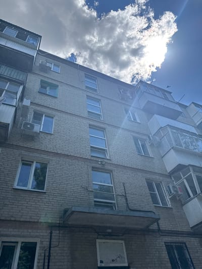 A low-angle, wide shot captures the facade of a multi-story brick apartment building under a bright, partially cloudy sky. The building, constructed of light-colored bricks, features numerous windows and balconies, many of which are enclosed with glass or siding. Air conditioning units are visible mounted on some of the exterior walls. The sky is a mix of blue sky and large, bright white clouds, with the sun partially obscured by one of the clouds, creating lens flare and a sunbeam effect. A small bird is silhouetted against the sky near the top of the building. The image appears to be taken in a residential area in the city of Azov, Russia, given the architectural style and common features of apartment buildings in the region.