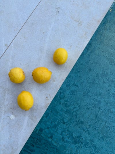 Four lemons are situated on the edge of a pool in Municipal Unit of Elliniko, Greece. The pool's edge is made of light-colored tiles, and the water is a turquoise color with reflections creating a rippled effect. The lemons are scattered across the tile surface, with varying shapes and textures visible. The scene is lit with natural light, highlighting the textures of both the lemons and the pool water.