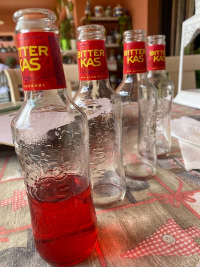 A close-up, eye-level shot shows four glass bottles of Bitter Kas, a Spanish aperitif, arranged diagonally on a table covered with a patterned tablecloth. The bottle in the foreground, on the left, is full of the dark red liquid, while the other three bottles are empty. The labels on the bottles are red with yellow lettering that reads "BITTER KAS" and "SIN ALCOHOL." The tablecloth has a festive pattern of red ribbons and bows. The background is blurred, but appears to be an outdoor setting with plants and furniture. The scene is set in Resinera-Voladilla, Spain.