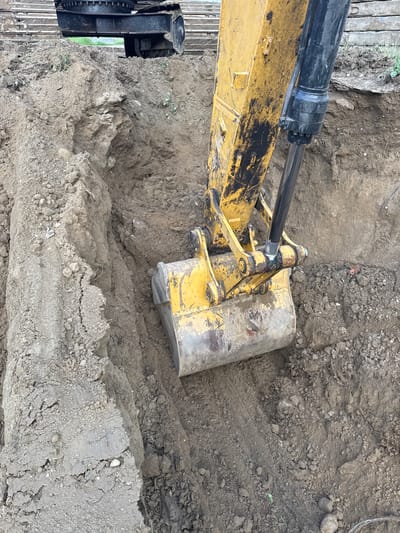 This media file captures an overhead view of an excavator bucket and its hydraulic arm, actively digging into a dirt trench. The excavator is predominantly yellow with black accents, showing signs of wear and tear. The scene is characterized by rough, excavated earth, with visible tire tracks imprinted in the soil. The ground is a mix of dirt and small rocks, suggesting an outdoor construction or digging site. The lighting suggests it is daytime, but the weather is not discernible. No people are visible in the frame. The focus is on the heavy machinery and the raw earth, emphasizing the physical act of excavation. The location context is Usinsk, Russia, but no specific visual cues within the image confirm this. There is no visible text in the media file.