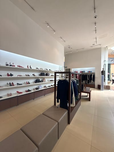 The image shows the interior of a retail store. The store appears to sell clothing, shoes, and accessories. Clothing is displayed on racks near the center of the image, while shoes are displayed on shelving units along the wall to the left. There is a seating area along the right side of the image, consisting of a row of upholstered blocks. The location is likely San Francisco Ocotlán, Mexico. The store appears well-lit with track lighting on the ceiling. No people are clearly visible in the foreground.