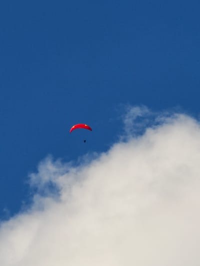 A paraglider is in flight against a bright blue sky. The paraglider's canopy is red with yellow accents, and the person is a dark silhouette beneath it. A large cloud formation is visible in the lower right portion of the image, partially obscuring the sky. The scene appears to be set in Zgorigrad, Bulgaria.