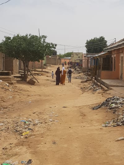 A dirt street in Dutsinma B, Nigeria, shows buildings along each side with garbage accumulating on the right side of the road. Two adults wearing traditional clothing walk along the road, accompanied by a small child. An adult wearing a backpack walks behind them. A tree is on the left side. Overhead wires span the street.