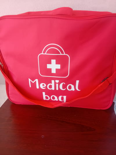 The image shows a bright red medical bag with white text that reads "Medical bag". A white cross symbol is also visible on the bag, enclosed in a stylized outline of a bag with handles. The medical bag is lying on its side, partially obscuring a dark red, wooden surface. A thin, red strap is attached to the bag and extends out of the frame. The background on the left is a textured, pale pink wall. The lighting suggests an indoor setting, possibly a home or office. The image is taken from a slightly elevated angle, looking down at the bag. There are no people visible in the image. The overall impression is that of a practical, easily identifiable medical supply bag.