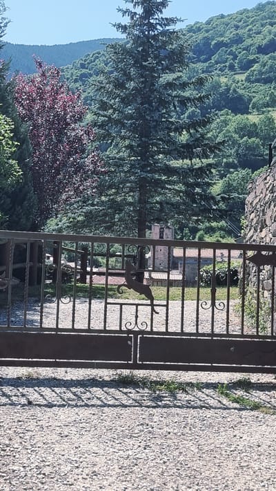 This image captures a view through a wrought iron gate in Setcases, Spain. The gate is adorned with a silhouette of a leaping deer. Beyond the gate, a textured gravel driveway leads towards a stone wall on the right. In the background, a landscape unfolds with lush green hills covered in dense foliage and trees. A traditional stone building with a red tiled roof and a bell tower is visible amongst the trees. The sky is clear and blue, suggesting a sunny day. The overall impression is one of a tranquil rural setting.