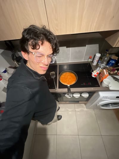 A person wearing glasses and a dark sweater is positioned in a kitchen. A frying pan with orange sauce is on a black stovetop. The countertop is cluttered with bottles, cans, and mugs. A washing machine is next to the stovetop. The floor is tiled.