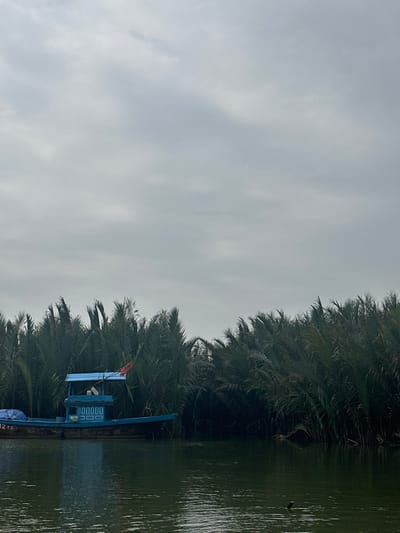 A partly cloudy sky hangs over a verdant landscape with water. Lush, dense palm trees line the water's edge. A blue, boat-like structure with a small flag is situated on the water. It appears to be docked alongside the vegetation, blending with the greenery. The scene evokes a sense of calm and tranquility, highlighting the natural beauty of a tropical environment.