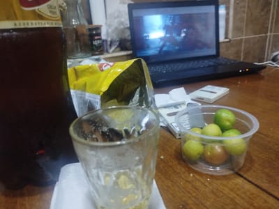 A still life photograph captures a cluttered wooden table. In the foreground, a dirty glass and a plastic container of small, round fruits, likely plums or apricots, are visible. Behind these items, a bag of chips, a pack of cigarettes, and a bottle of dark liquid, possibly soda or beer, are placed. Further back, a laptop displays a bright, blurred image, and in the background, a small jar with a dark substance and a glass bottle are also present. The scene is set indoors, with tiled walls and a wooden surface suggesting a casual setting. The image lacks any human presence. The provided context suggests the location is Sabunchu, Azerbaijan.