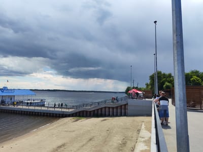 The image depicts a waterfront scene in Volsk, Russia, on a cloudy day. In the foreground, a sandy embankment slopes down to the dark water of a wide river. To the left, a long pier extends into the water, featuring a building with a bright blue roof and the Cyrillic inscription "ВОЛЖСК" (Volzhsk). Several people are visible on the pier, near the building and along its edge. A wide, paved walkway with a metal railing occupies the right side of the image, leading towards the river. A group of people are standing on this walkway, facing the water. In the background, the river stretches towards a tree-lined horizon under a dramatic sky filled with dark, heavy clouds, with hints of sunlight breaking through in some areas. Several lampposts are visible along the walkway and in the distance.