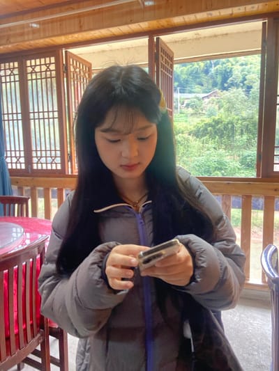 A young woman with black hair and bangs wearing a gray puffer jacket holds a cell phone in her hands. She looks down at the device. The background reveals a rustic wooden structure with windows overlooking a green, hilly landscape. A red table and chairs are visible in the foreground, suggesting an indoor setting. The image is oriented vertically.