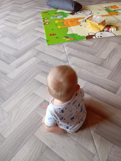 A baby is sitting on a light-colored wooden floor in an indoor setting. The baby is facing away from the camera, with their back and head visible. They are wearing a white onesie with a pattern of blue cars and stars. To the right of the baby, on the floor, is a colorful playmat with cartoon animal illustrations. On the playmat, there is a small yellow patterned bag and a few other objects, including what appears to be a toy. A gray pillow is partially visible in the upper portion of the image. The floor has a herringbone wood pattern. The scene is likely a home environment in the city of Vysokaya Gora, Russia.