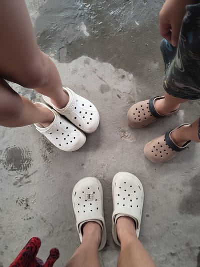 The image shows a top-down view of the feet of at least three individuals standing on a wet concrete surface, possibly an outdoor area in Dustabad (Soldatsky), Uzbekistan. Two pairs of white Crocs are visible, one pair is worn by a person with their legs crossed. Another pair of tan Crocs is worn by another individual. The wet concrete surface suggests it may have recently rained or been washed. Footprints are visible in the wet concrete. One person is wearing a red and black patterned glove on one hand, which is also visible in the frame. The overall scene is a casual, outdoor setting.