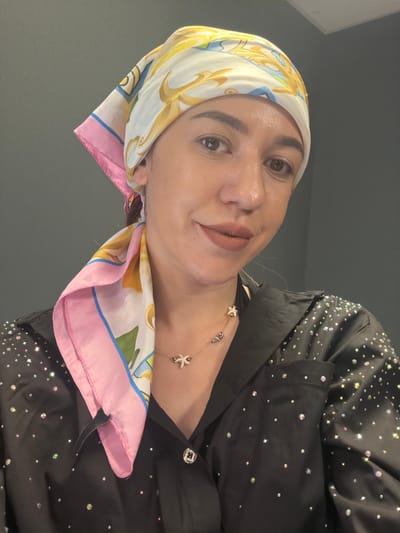 A woman is captured in a close-up, head-and-shoulders portrait. She is wearing a brightly patterned silk headscarf, primarily white with yellow, blue, and pink accents, tied loosely around her head. Her dark hair is visible beneath the scarf. She has brown eyes, well-defined eyebrows, and is wearing matte brown lipstick. A delicate necklace with small star and bead charms rests on her collarbone. Her clothing is a black, button-down shirt adorned with numerous small, iridescent rhinestones scattered across the fabric, particularly concentrated on the shoulders and upper chest. The background is a plain, dark gray wall. The scene appears to be an indoor setting, possibly a room in a residential or hotel property in Hisarya, Bulgaria.