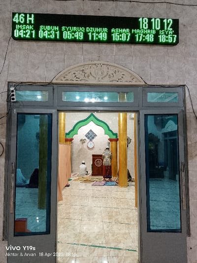 The image shows a view through the doorway of a mosque located in Kecamatan Kroya, Indonesia, with coordinates (latitude: -6.423, longitude: 107.996). The doorway is flanked by two closed doors. The interior of the mosque is visible through the open doorway. Two individuals are present, both wearing traditional Islamic attire. The person on the left is seated on a prayer mat, while the person on the right is standing and holding a prayer book. The floor is tiled, and there are three pillars on the right side of the frame. A clock is visible on the wall behind the standing individual. An electronic sign above the doorway displays prayer times for the day.