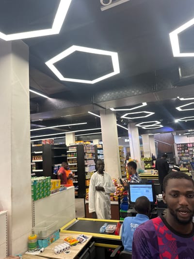 The image displays the interior of a brightly lit supermarket in Babban Giji, Nigeria, characterized by a black ceiling with numerous hexagonal and linear LED light fixtures. The foreground on the right features a young adult male with a beard, wearing a purple patterned shirt, looking to the right. Directly behind a checkout counter in the mid-ground, a man in a light blue "SUF" branded shirt sits, operating a computer monitor displaying a retail interface. The counter holds rolls of receipt paper, snack bags, two plastic water bottles, and a plastic container with red contents. To the left of the counter, stacked green-packaged cans are visible on an adjacent white counter surface.

An adult male in a long white traditional garment stands upright to the left of the checkout area, facing the viewer. Behind him, a young adult male in a blue plaid shirt is engrossed with a smartphone. The background reveals multiple aisles lined with dark shelving units stocked with various retail products. A male in an orange shirt is positioned near these shelves on the left. Further to the right in the background, another counter with large fruits (possibly watermelons) is visible, along with additional shoppers and a sign partially displaying "SUPPLEMENTS." The scene depicts a modern, active retail environment.