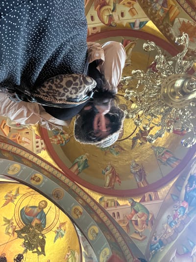 The image shows a person looking up at a domed ceiling adorned with religious frescoes and a large, ornate chandelier. The frescoes depict figures in religious attire, possibly biblical scenes, against a gold background. The person is wearing a black polka-dot dress and has a leopard-print bag slung over their shoulder. The lighting suggests an interior space, likely a church or religious building, with sunlight filtering in. The scene is oriented upside down. The location is stated to be the city of Migdal, Israel, which is home to the Migdal Synagogue, a reconstructed ancient synagogue with similar decorative elements.