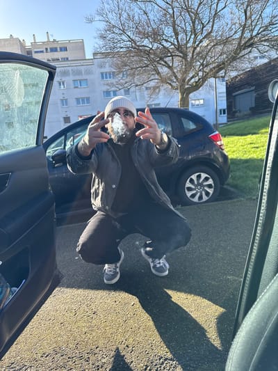 An adult male is captured in a crouched pose within a bright outdoor setting, consistent with a residential area in Hérouville-Saint-Clair, France. The subject, positioned between two open car doors, wears a light-colored beanie, dark sunglasses, a dark jacket over a dark shirt, dark trousers, and light grey/white athletic shoes. He has a dark beard and mustache. Both hands are raised, featuring an extended index and pinky finger with the middle and ring fingers bent. A metallic watch is visible on his left wrist. The subject is actively exhaling a dense cloud of white vapor or smoke directly from his mouth, partially obscuring his lower face.
In the midground, a dark-colored compact hatchback vehicle is parked on asphalt pavement. Behind it, a patch of green grass is visible, along with a large, bare deciduous tree. The background features several multi-story, light-colored residential buildings with multiple windows, typical of apartment blocks. To the far right, a darker, possibly wooden, building with a large door is present. The scene is illuminated by bright sunlight, casting sharp shadows from the subject and vehicles onto the asphalt ground. The photograph appears to be taken from inside a vehicle, with its open driver's side door visible on the left and a portion of the passenger side interior on the right.
