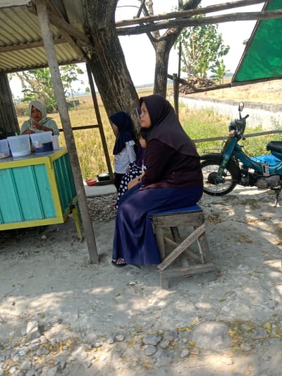 In Kemamang, Indonesia, a side view captures a simple roadside stall with a woman in a light green hijab serving customers from behind a teal and yellow counter laden with clear plastic containers of drinks. To the right, another woman in a dark purple hijab and a long, dark blue skirt sits on a wooden stool, facing away from the viewer. Behind her, a child in a dark blue hijab and a white shirt with polka dot pants stands. The scene unfolds under a rustic, open-air shelter with a corrugated metal roof supported by wooden beams. A large tree with textured bark is integrated into the structure, casting shadows on the dusty ground littered with dry leaves and small stones. Beyond the stall, a field of dry, light brown vegetation stretches out, bordered by a low concrete wall. A turquoise motorcycle with a basket is parked to the right of the stall, its handlebars and front wheel visible. The overall atmosphere suggests a humble, everyday transaction taking place in a rural or semi-rural setting.