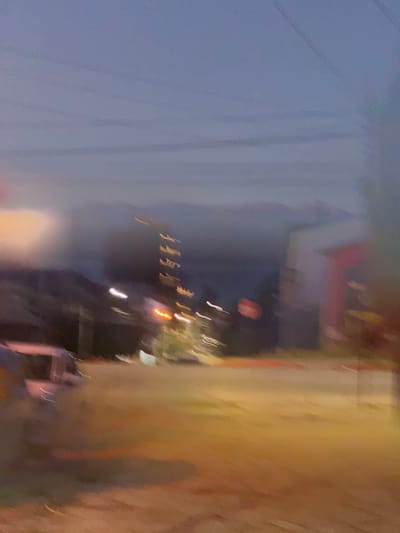 Blurred and distorted image of a street scene. Visible elements include buildings, streetlights, and utility wires against a dusky sky. The image is rotated, obscuring detail and creating a sense of motion or instability. Color tones vary from light blues and grays to yellows and oranges.
