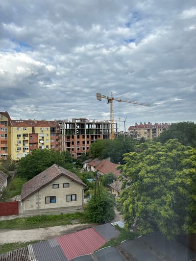 The image depicts an urban scene in Subotica, Serbia, characterized by a mix of existing residential buildings and ongoing construction. In the foreground, a low-rise house with a tiled roof and several windows is visible, showing some signs of wear and graffiti on its exterior walls. Adjacent to this house is a red metal gate and a patch of green grass. Further into the scene, to the right, a large, leafy tree dominates the view.
Behind these elements, a multi-story apartment building, painted in yellow and terracotta colors, stands with its numerous windows and balconies. To the right of this building, a large construction site is evident. A tall yellow construction crane is prominently positioned, with a partially built concrete structure of a new building behind it. More residential buildings with red tiled roofs can be seen in the background, suggesting a developed urban area. The sky is overcast with dramatic, grey clouds, contributing to a subdued lighting atmosphere. The presence of construction indicates urban development and expansion within the city.