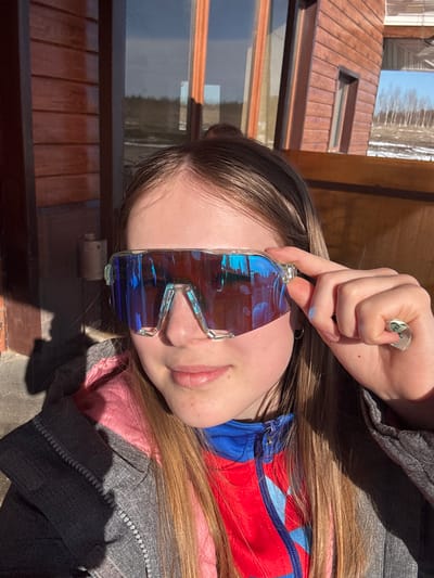 A young woman is outside in daylight near a building with brown horizontal siding and a brown roof, possibly in the vicinity of Gory, Russia. She is wearing a gray zip-up jacket over a red and blue zip-up top, and she is holding a pair of blue-tinted, futuristic-looking sunglasses in front of her face. A folded piece of paper, possibly money, is held near her forehead. The image is oriented sideways.