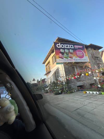 The image captures a view from a car window, looking out at a street scene in Kresna, Bulgaria. In the foreground, a fluffy keychain shaped like a green and white bird hangs from the car's interior. The main focus of the scene is a building with a large billboard advertising "dozza ENERGY SHOT." The billboard features images of three colorful bottles and text in Bulgarian that translates to "pure energy for your day..."
The building itself appears to be aged, with visible brickwork and a sloping roof. A canopy extends from the front of the building, and numerous potted plants, including red and pink flowers, are arranged along the facade and on the ground. A sidewalk made of grey square tiles stretches across the foreground and midground. In the distance, a triangular traffic sign is visible, along with glimpses of other buildings and a clear blue sky. The overall impression is of a sunny day in a small town.