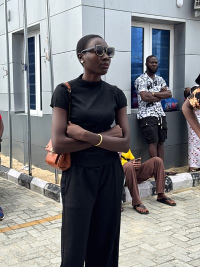 The image shows a group of people in Ojomu, Nigeria. A person in the foreground wears black clothing and sunglasses, with a small brown purse. Behind them, another person lies on what appears to be a sidewalk or ledge with their arms crossed, wearing a patterned shirt and black shorts. Another individual wearing brown pants sits near them. The environment suggests an urban setting with a building in the background. The ground appears to be paved with gray bricks and has a yellow stripe on it.