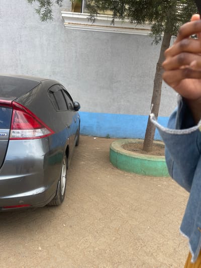 This outdoor scene is situated in the Hospital ward, Kenya. A dark grey, two-box passenger vehicle, identified by visible "Hybrid" badging and resembling a Honda Insight, is parked on a light brown patterned ground surface, positioned on the left side of the frame with its rear facing the viewer. The vehicle exhibits a layer of dust. In the background, a building wall features an upper section of light grey textured stucco and a lower section of vibrant blue paint. A white decorative architectural trim is present above the grey wall. To the right of the vehicle, a slender tree with sparse green foliage grows from a circular, green-painted concrete planter. The immediate foreground on the right displays a person's right hand, clasped, and part of a denim jacket sleeve over a yellow undergarment, partially obscuring the lower right corner of the frame.