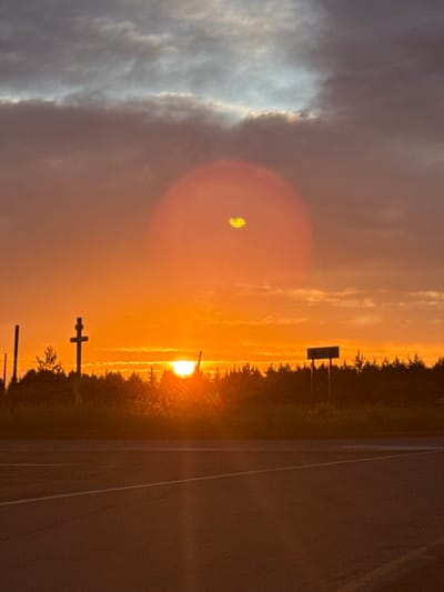 The image captures a sunset over a rural landscape, with a road in the foreground. The sky is filled with dramatic clouds, ranging from dark grey to vibrant orange and yellow, with the sun appearing as a bright orange disc near the horizon. A lens flare, a circular orange glow with a yellowish center, is visible in the upper portion of the sky.
In the middle ground, a silhouetted treeline stretches across the scene, with a few distinct objects protruding above it. To the left of the center, a dark, cross-shaped structure stands prominently. To its right, a tall, thin pole is visible. Further to the right, a rectangular sign mounted on two posts stands out against the treeline and sunset. The sun is partially obscured by the trees on the right side of the image.
The foreground consists of an asphalt road with white lane markings, suggesting it's a highway or a significant road. The overall impression is one of a remote, serene setting bathed in the warm light of a setting sun.
This scene is likely located in the vicinity of Гольяны, Russia, a village in the Tver Oblast, known for its rural character and natural landscapes, fitting the depiction of a sunset over a treeline and road. The presence of the cross structure might indicate a religious or memorial site, common in rural Russian landscapes.