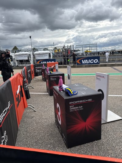 An outdoor scene depicts a preparatory area at the Sachsenring circuit in Hohenstein-Ernstthal, Germany, under an overcast sky. In the foreground, black and orange barriers with "PORSCHE SIXT CARRERA CUP" branding line the left side of an asphalt surface. Three black rectangular boxes, marked "3," "2," and "1" respectively, are arranged in a row towards the background, each featuring "PORSCHE SIXT CARRERA CUP DEUTSCHLAND" text and a red graphic. The box marked "3" has a pink water bottle, a white cloth, and a blue and yellow cap on top. Boxes "2" and "1" each have a pink water bottle and a cap (only one visible on box 2). Adjacent to box "1" is a tall white sign with a black "0." To the far left, a person wearing dark attire holds a camera, facing right. Beyond a low concrete barrier, two individuals are visible; one in dark clothing gestures with both arms, while another in white racing overalls stands on the barrier looking right. In the background, "MANTHEY EMA" branded service vehicles and a "VAICO" banner are visible, with distant spectators behind fencing.