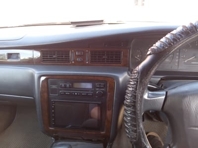 The image shows the interior of a car's dashboard, specifically the area around the steering wheel, air conditioning controls, and a display screen. The dashboard features wood-grain trim and black plastic components. A leather steering wheel cover with a textured pattern is visible. The car appears to be parked, as indicated by the lack of movement blur and the static nature of the dashboard elements. The lighting suggests it is daytime, with light filtering in from outside the vehicle. There is no information in the image to suggest it is located in Aldan, Russia.