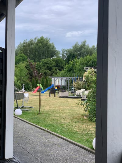 The image depicts a backyard scene in Rewal, Poland. The foreground shows a white wooden beam and a dark railing, suggesting a porch or balcony. Beyond this, a green lawn stretches out, dotted with trees and bushes. In the middle ground, a playground structure with red, green, and blue slides is visible. A hammock with a white seat is also present, along with a small tree with dark leaves. Further back, more trees form a dense green backdrop. The sky is cloudy, indicating a potentially overcast day. The overall setting appears to be a residential backyard, likely in a suburban or rural area, given the abundance of greenery. No people are visible in the image, and no text is present. The scene conveys a sense of tranquility and leisure, with the playground suggesting a family-friendly environment. The time of day appears to be daytime.