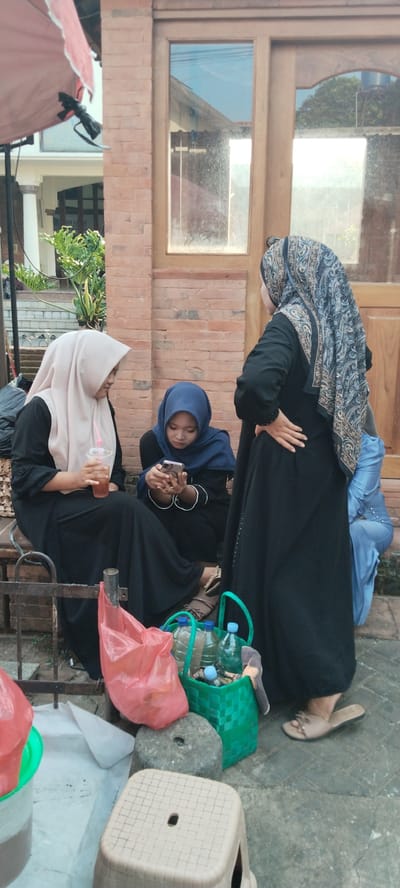 Three women, dressed in hijabs and long black dresses, are gathered outdoors in front of a brick building with a wooden door and windows. The woman on the left is holding a drink in a clear plastic cup with a straw and is wearing a light pink hijab. The woman in the center is wearing a dark blue hijab and is looking down at her phone. The woman on the right is wearing a patterned hijab and has her hand on her hip, facing away from the camera. In the foreground, there are several items on the ground, including a red plastic bag, a green woven basket containing several plastic bottles of water, and a small plastic stool. The ground is paved with stone. The image appears to have been taken during the daytime, with natural light illuminating the scene. The setting is the city of Kauman, Indonesia.