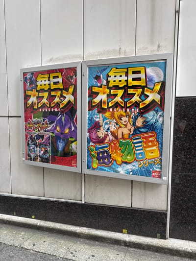 Two framed posters are mounted side-by-side on a textured, off-white wall. The poster on the left features anime-style characters and graphics with bold yellow Japanese text across the top. Prominently displayed in the lower half is a large, purple robot with glowing yellow eyes. The poster on the right also has bold yellow Japanese text across the top and features several anime-style female characters with colorful hair. Below the characters, a title in colorful, three-dimensional Japanese text is visible, along with the word "シリーズ" (series) in smaller text. Both posters have silver frames and appear to be advertisements, likely for pachinko or slot machines, given the small white insert with Japanese text and a number "13" in the bottom right corner of the right poster. The wall itself is segmented by vertical lines and shows some signs of dirt or staining. Below the wall is a dark, speckled granite base that extends to the ground. The ground in front of the posters is a concrete sidewalk with some wear and tear, and a curb separates it from a darker asphalt street. The overall scene suggests an urban environment, consistent with Shinjuku, Japan, where such advertisements are common.