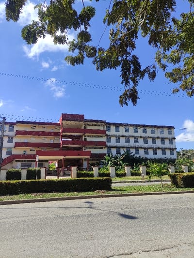 The image depicts a multi-story concrete building under a bright blue sky with scattered white clouds, situated in Ciego de Ávila, Cuba. Dark green tree branches with dense foliage frame the upper portion of the sky, with thin dark lines, likely wires, visible horizontally across the sky.
The building, appearing institutional or residential, features a light yellow or cream facade with prominent deep red exterior elements, particularly on a stepped series of balconies and stairwells on its left side. The right side of the building shows a grid pattern of numerous windows, many appearing darkened or covered. Signs of wear and discoloration are visible on the building's exterior.
In the mid-ground, a dark green, manicured hedge runs parallel to a paved asphalt road in the foreground. Behind the hedge, a grassy strip contains a series of light-colored, square-sectioned pillars, between which a metal gate is partially visible on the left. Lush green vegetation, including what resembles banana plants, grows immediately behind the pillars and around the building's base. A small tree with sparse green leaves stands in the grassy area on the right.
The asphalt road in the foreground is dark grey, showing surface texture and minor cracks. A shadow, likely from a light pole, is cast onto the road in the lower center. No individuals, vehicles, or specific actions are visible within the scene. The environment appears calm under daylight conditions.