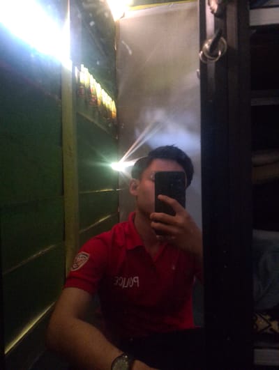 This is a mirror selfie of a young man, likely taken indoors. He is wearing a red polo shirt with "POLICE" written on it in white lettering, and a patch on his left sleeve that appears to be a police badge. He is holding a black smartphone to take the picture, covering most of his face. His right arm and shoulder are visible, as is a portion of his left arm with the police patch. He is wearing a watch on his left wrist.

The setting appears to be a narrow hallway or an elevator. To the left, there's a green wall and some bright lights emanating from a fixture above and to the left of the man, creating lens flare effects in the image. The light suggests it might be daytime, or at least a brightly lit interior space. On the right side of the frame, a dark vertical structure, possibly a door frame or a closet door, is visible. The mirror's surface is reflective, showing the man and the environment around him. The overall impression is casual, possibly taken in a backstage area or a private space.
