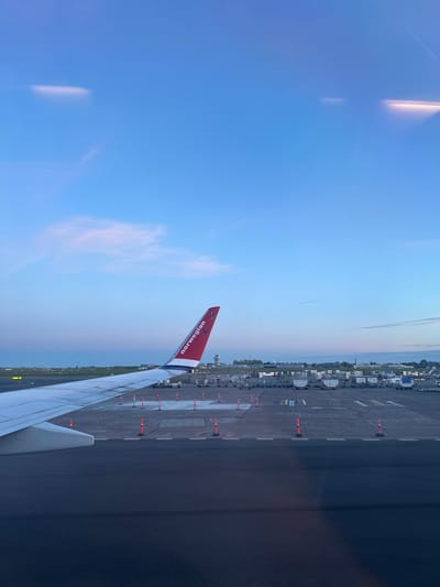 The image depicts the wing of a Norwegian Air Shuttle airplane, viewed from a passenger window. The red winglet displays the airline's logo. Beyond the wing, an airport tarmac is visible, marked with orange cones and painted lines. Further in the distance, there are buildings and possible airport infrastructure. The sky is a gradient of blue, suggesting either sunrise or sunset. The scene is likely taken at Copenhagen Airport (CPH), located in Kastrup, Denmark.