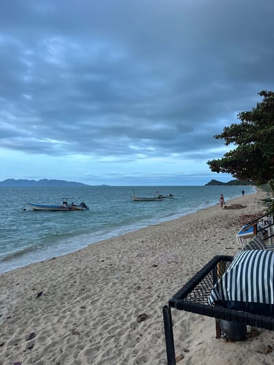 The image depicts a serene beach scene with a person and a boat. The main subject is a person standing on the sandy shore, looking out towards the ocean. A traditional longtail boat is anchored in the shallow water near the shore, with its distinctive wooden hull and engine visible. The setting is a tropical beach, with fine sand and gentle waves lapping at the shore.
The time of day appears to be late afternoon or early evening, as indicated by the soft, diffused light and the slightly overcast sky. The weather is calm and pleasant, with a light breeze suggested by the movement of the water. The location cues suggest a Thai beach, possibly in Baan Thong Po, with the characteristic longtail boat and the lush green foliage from a nearby tree.
The overall mood is tranquil and relaxed, inviting viewers to imagine themselves at this peaceful coastal retreat. There is no visible text in the image.