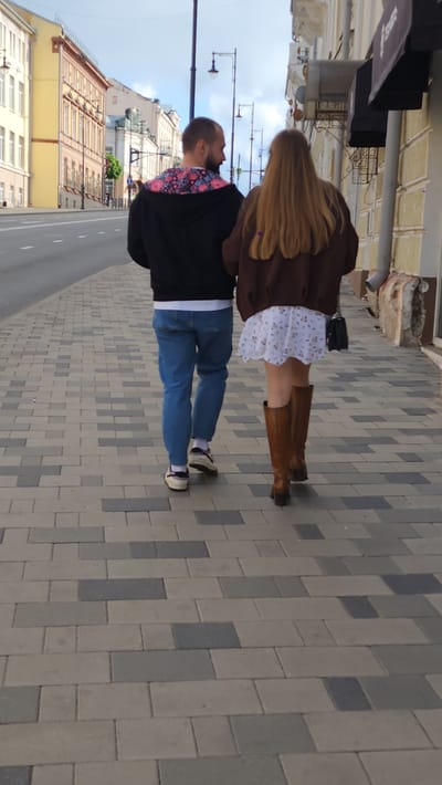 A man and a woman are walking side-by-side on a paved sidewalk in what appears to be a city in Russia. The woman is dressed in a white dress with a floral print, brown knee-high boots, and a brown jacket, and has long blonde hair. The man is wearing blue jeans, a black jacket with a colorful patterned collar, and white socks with sneakers. They are on a sidewalk made of gray and brown rectangular paving stones. To their left is a street with yellow buildings on the other side, and lamp posts line the sidewalk. The buildings have classic European architectural styles with multiple windows. The location is identified as Smolensk, Russia.