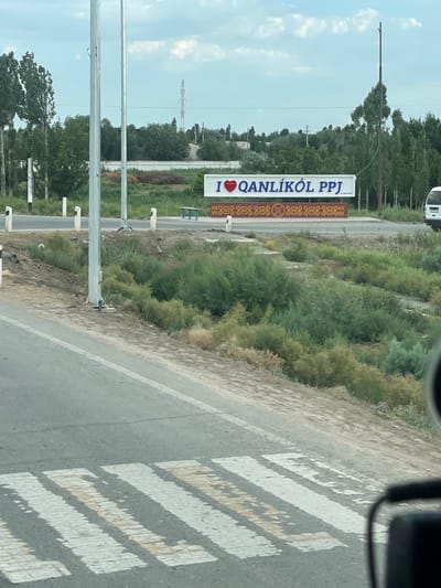 This media file depicts a scene in Qanlikul, Uzbekistan. The primary subject is a roadside sign with bold white text that reads "I ❤️ QANLİKÖL PPJ". The sign also features decorative elements on its right side. The scene appears to be outdoors, likely during the day, with a slightly overcast sky visible. Trees and dense vegetation line the roadside, suggesting a rural or semi-urban setting. A paved road with a pedestrian crossing marked by white stripes is also visible, indicating a public area. The perspective suggests the image was taken from within a vehicle. There are no people clearly visible in the frame. The overall impression is one of a welcoming sign for the region of Qanlikul.