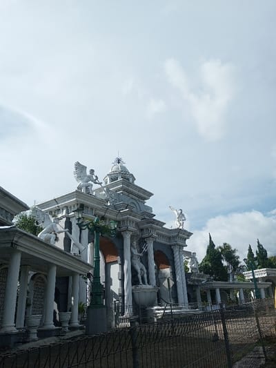 A grandiose structure with a classical theme features multiple statues of winged figures and muscular men with tridents. White pillars and arched entrances adorn the building, topped with a dome-like structure. A tall, green lamppost stands in front. A black chain-link fence runs along the bottom of the frame. The sky is partly cloudy. Green trees are visible in the background.