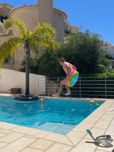 A medium, eye-level shot reveals a person mid-leap into a swimming pool at a location identified as el Poble Nou de Benitatxell / Benitachell, Spain. The person, a muscular male, is airborne with a multi-colored inflatable seat pad positioned around his waist.
The pool, filled with clear blue water, features a large black tire floating in it, with two multi-colored balls present. The poolside is paved with beige-colored tiles. A full-face snorkel mask rests on the pool deck, partially out of frame. A white metal fence separates the pool area from a lush green shrubbery, behind which the exterior of a building can be seen. A palm tree stands at the edge of the pool area. The sky is clear and blue.