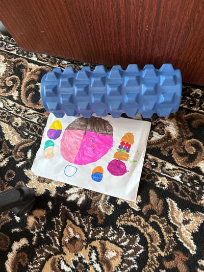 A blue foam roller is placed on a patterned carpet next to a drawing on white paper. The drawing depicts several colorful items, including what appear to be ice cream cones and burgers. The carpet has a dark base color with intricate patterns in shades of brown, beige, and white. In the background, a portion of a wooden surface, possibly furniture, is visible. A black object, potentially a chair leg, is partially visible in the lower left corner. The scene appears to be indoors, likely in a residential setting. No specific location in the city of Urazovo, Russia, can be determined from the image alone.