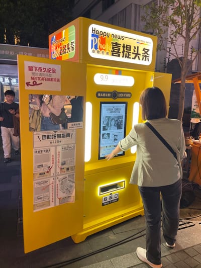 The image shows a yellow photo booth, located on a street in Hangzhou Shi, China at the coordinates 30.263 latitude and 120.156 longitude. The photo booth is labeled "Happy News Photo Booth" and is adorned with Chinese and English text. A woman is interacting with the machine, possibly paying for or selecting a photo option. The screen displays a headline for a news article, visible behind the woman. The photo booth is situated on the sidewalk next to a building. The pavement is clean and a black cable snakes across the ground. The photo booth has a large yellow door open, revealing a sign with Chinese characters. To the left of the photo booth, a man stands with a backpack on, looking down at his phone. Behind the booth, the street is dimly lit, suggesting it is nighttime. A sign hanging above the booth's door reads "NestCafe." To the left of the photo booth, a sign that says "Welcome to Nemcafe" is visible. The scene suggests a bustling city environment.