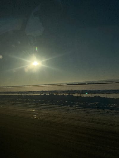The image depicts a vast, snow-covered winter landscape in Озерный, Russia, viewed from a slightly elevated perspective, likely from inside a vehicle.
**Foreground**: The lower third of the frame is occupied by a dark, textured surface, possibly a road or a frozen body of water, showing longitudinal streaks and reflective patches consistent with melting ice or water. Visible reflections suggest the image was captured through a window.
**Middle Ground**: Immediately beyond the foreground, a substantial bank of uneven, piled snow runs horizontally across the scene. Behind this snow bank, an expansive, flat area is uniformly covered in snow, stretching into the distance. Sparse, faint vertical elements, possibly fence posts or distant structures, are discernible across this snow-covered plain.
**Background**: The snow-covered plain extends to the horizon, where a hazy, dark line of distant hills or low mountains is visible.
**Sky**: The sky transitions from a bright, hazy white-blue around the sun to a darker blue-grey towards the top of the frame.
**Sun**: A prominent, intensely bright sun, positioned in the upper-left quadrant, emits strong starburst light rays and exhibits significant lens flare, including a distinct green-blue artifact. Its low position indicates either early morning or late afternoon.
No people, animals, or active subjects are visible. The environment is cold, clear, and rural or semi-rural, characterized by extensive snow cover and bright, low-angle sunlight.