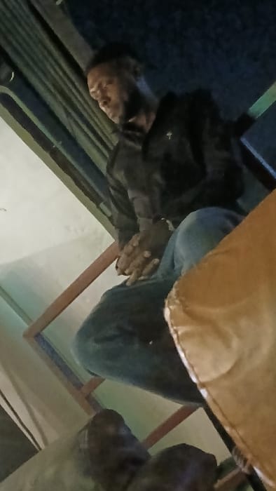 An individual seated, clad in a dark jacket, blue jeans, and wearing a watch on their left wrist, rests their hands together. A tan cloth drapes partially in the foreground. The setting includes a wooden chair, with a beige wall visible behind. The perspective is somewhat askew, casting an unusual angle.